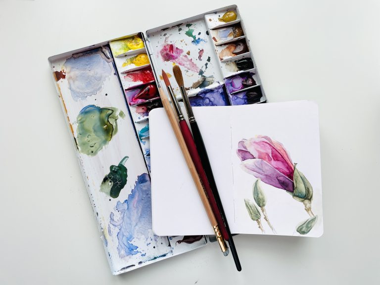 Watercolor paint palette with brushes and notebook containing a purple painted flower.