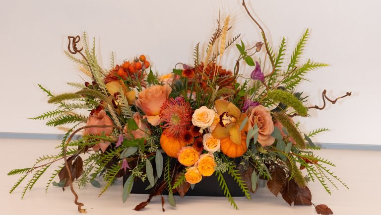 A fall themed centerpiece with flowers in ranges of yellow and orange set against a white backdrop.