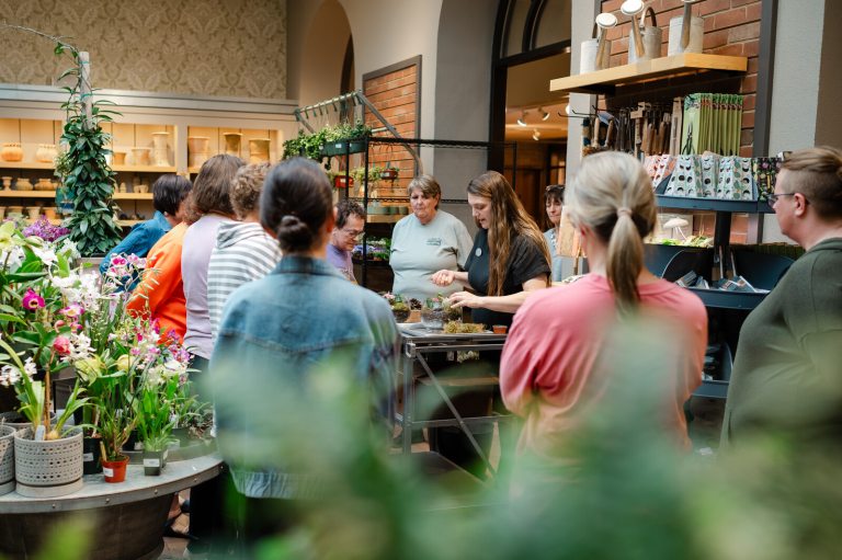 People in the Longwood Garden Shop, surrounding a table, watching someone pot a plant.