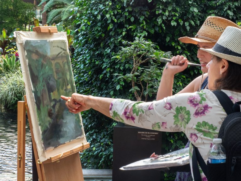 Two people looking and pointing at a painting in progress on an easel in a vegetated area.