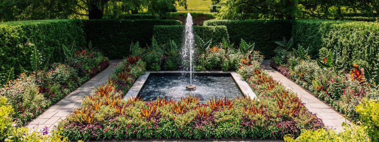 A single fountain jet rises above a square pool surrounded by colorful flowers and foliage.