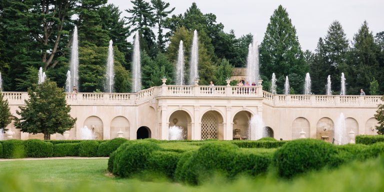 Fountains shoot up in straight lines from a white stone facade in a garden with bright green boxwood shrubs in the foreground