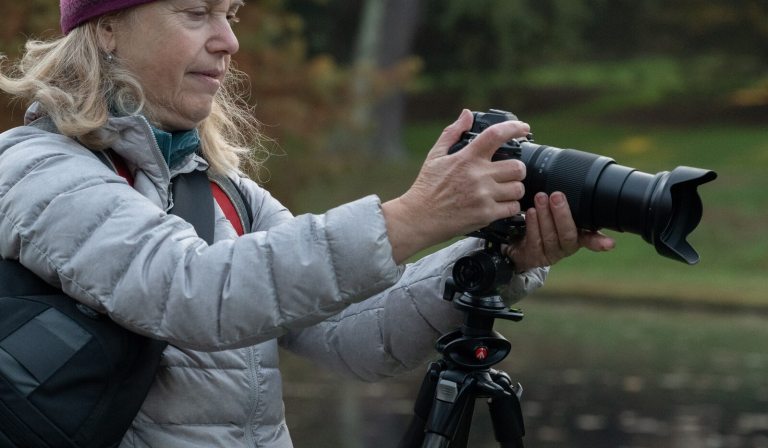 A person in a long coat and hat, taking a picture with a camera on a tripod.