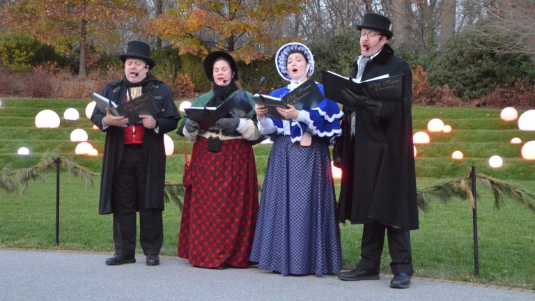 Four singers dressed in old-fashioned Dickens-era clothing and holding black songbooks, stand and sing in front of a lawn decorated with globes of white light.