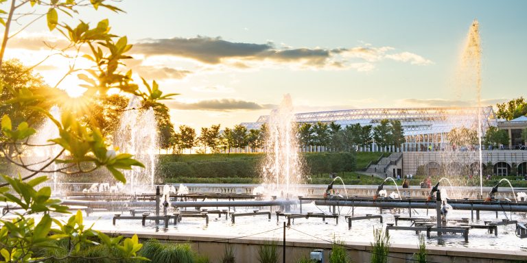 Sunset-lit fountains in foreground, with large conservatory complex in background.