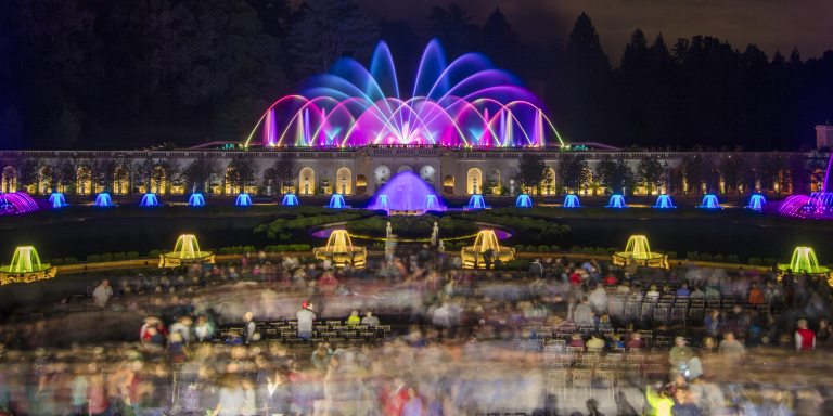 A wide shot of colorful fountains, with a blurred image of audience members talking, walking, and taking pictures in the foreground.