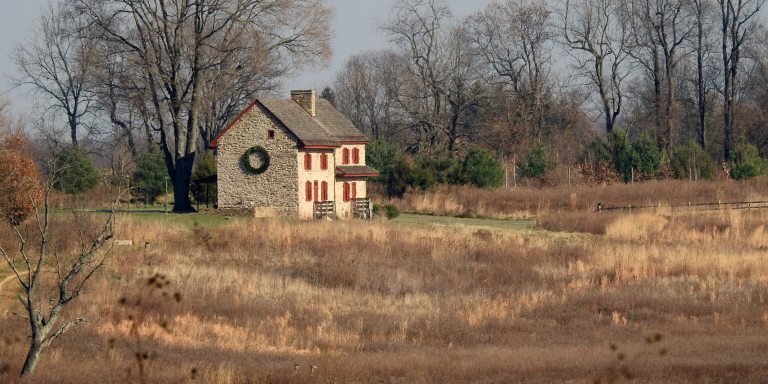 A two-story stone farmhouse with red shutters bears a green wreath on one wall, amid a late fall meadow.