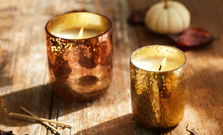 Two lit candles in metallic, mottled glass holders on a rustic wooden table, next to a small white pumpkin and fallen leaves.
