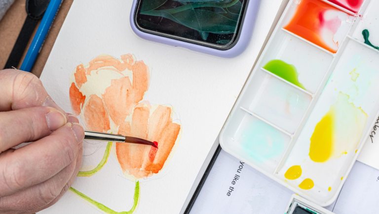 Close-up view of an artist painting an orange watercolor flower on paper, with a palette of vibrant paints and a smartphone visible nearby.