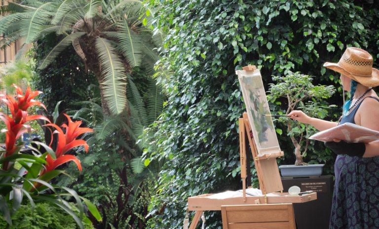 An artist wearing a straw hat paints a bonsai tree on an easel while surrounded by lush tropical plants and red bromeliads in a conservatory.