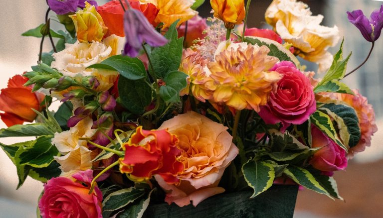 A close-up of a colorful, multi-layered bouquet with roses, tulips, and various blooms in shades of pink, yellow, orange, and purple.