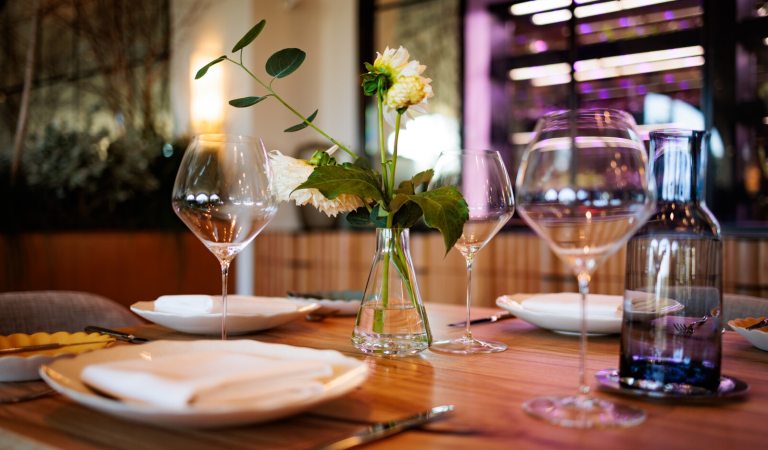 Set dining table with wine glasses, white plates, a napkin, a water bottle, and a small vase with a single white flower, suggesting a fine dining atmosphere.