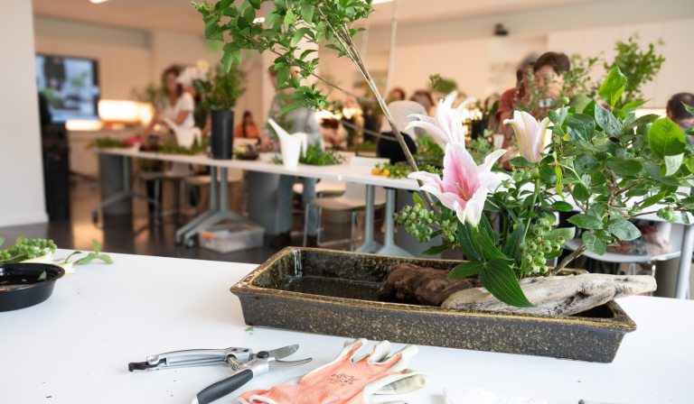 An indoor floral design workshop with an arrangement in the foreground and other participants working at tables in the blurred background.