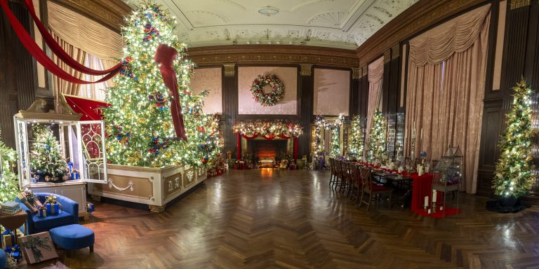 A formal room with polished wood floor, fabric walls, and ornate ceiling is decorated for Christmas with lit and decorated trees, a large red bow, a table set for dinner, and a fireplace with wreath, garland, and gifts.