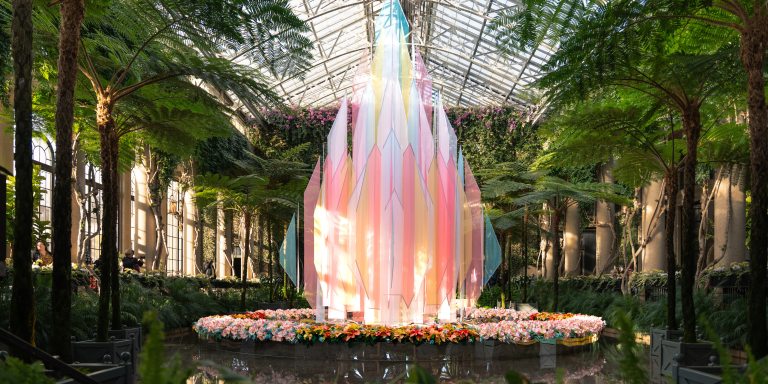 Geometric cloth panels in sheer pastel colors combine to form a diamond-shaped Christmas tree amid potted Christmas plants in a lush indoor conservatory setting.