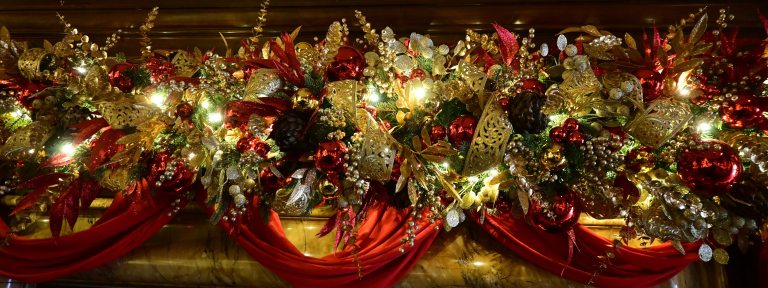 Closeup of a mantel decorated with swags of red cloth and an ornate garland in tones of red, gold, and silver.