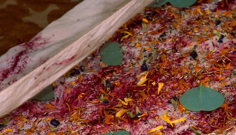 The fabric is covered with a dense layer of dried organic matter, including deep pink or purple petals, bright orange-yellow flower fragments, and small green eucalyptus leaves.