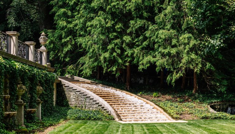 A garden featuring an ivy covered stone wall, with a staircase of rushing water leading to a manicured lawn. 