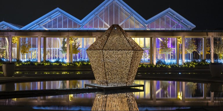 A giant metal lantern is reflected in a pool of water at night, backed by a glass conservatory lit from within in hues of blue and purple.