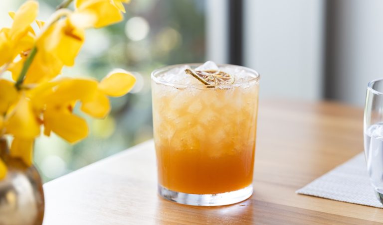 A peach drink, in a highball glass, set ona light wood table, with a vase of yellow flowers.
