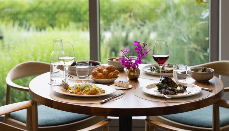 A table in a restuarant with plates of food and beverages, untouched, overlooking a green outdoor space.