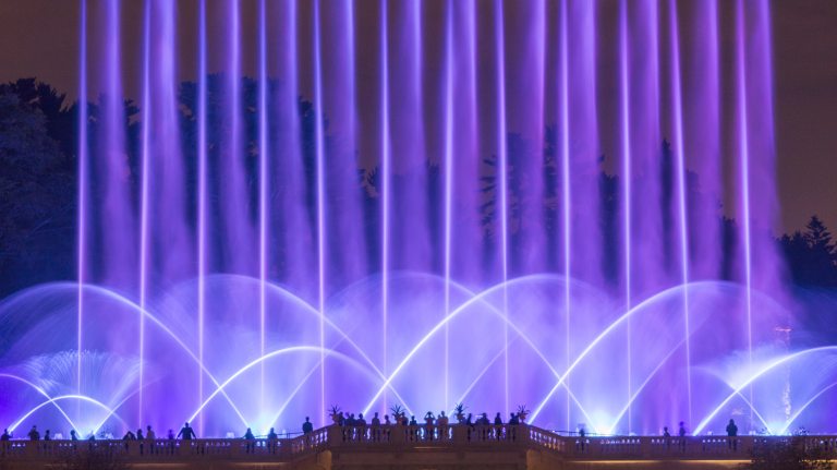 Fans and jets of fountain water bathed in lavender light rise above silhouettes of guests on a stone observation balcony.