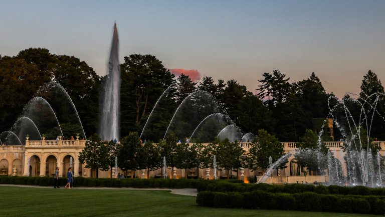 Wide view of a fountain garden at dusk, against the silhouette of trees and a clear deep blue sky.