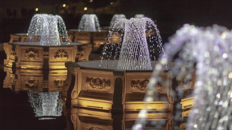 Basketweave fountains in octagonal basins lit by sparkling white light against a black sky.
