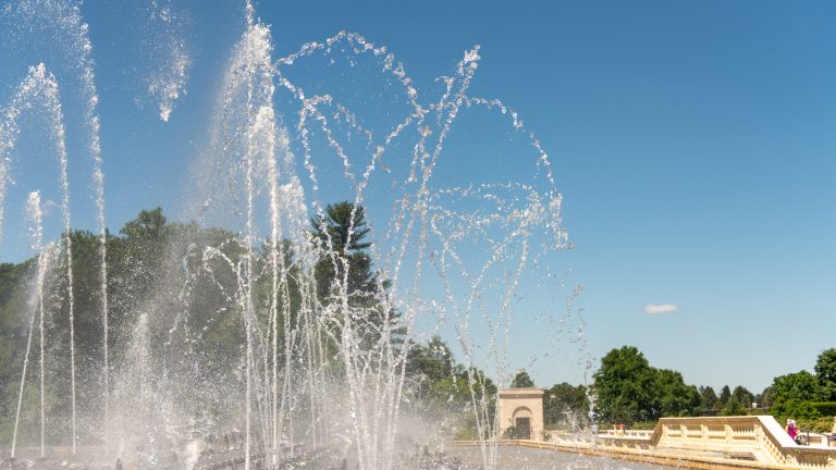Fountain jets of dancing water rise up from a stone basin against a cloudless blue sky.