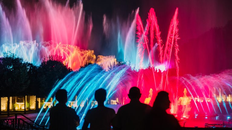 Four foreground silhouettes watch a nighttime fountain show in red, blue, and gold