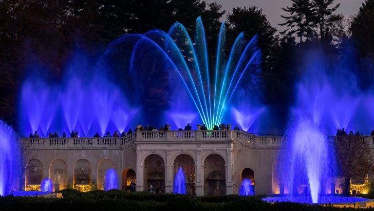 Fountains of water rise in hues of blue from the front and top of a stone facade.