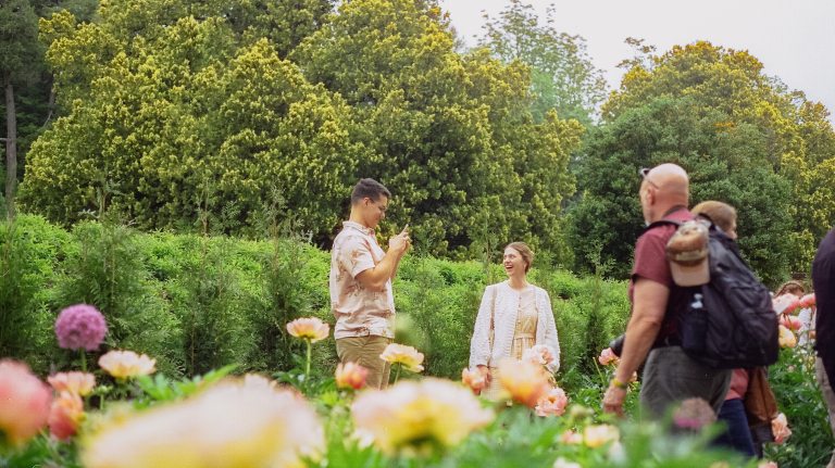 A guest with a smartphone captures the broad smile of another guest amid the blooms of a peony garden.