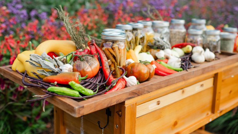 Closeup of wooden cart laden with fresh vegetables and filled mason jars, against a garden backdrop of red and purple blossoms.