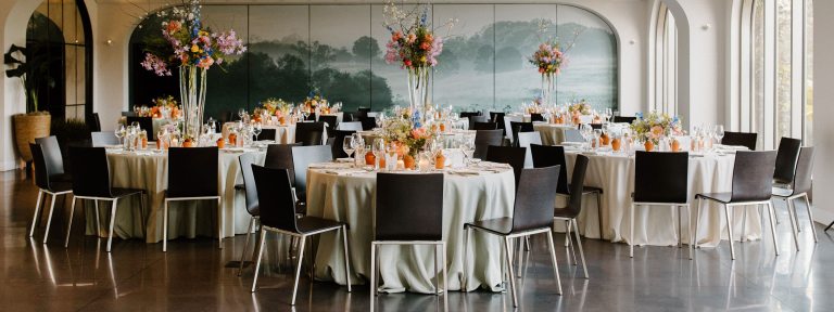 Seven tables are set for guests to dine at with linens, plates, and tall floral arrangements in a room with shiny black floors and a green mural on the back wall. 