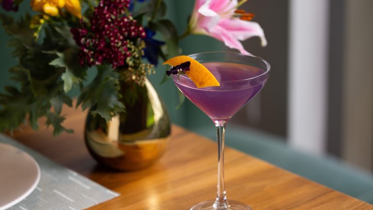 A long-stem cocktail glass holds a purple drink garnished with a flower and orange peel, with a gold-colored vase of summer flowers in the left background.