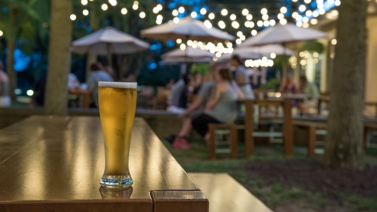 A glass of beer sits on the edge of a picnic table, against a background of guests at tables under party lights and table umbrellas.