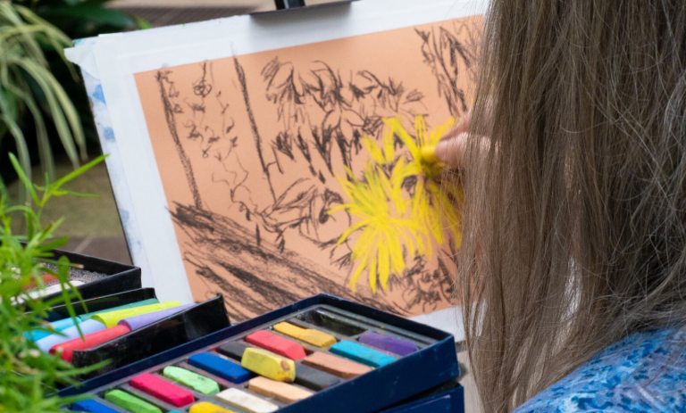 A close-up view over the shoulder of an individual using a bright yellow pastel stick to draw flowers on orange paper, with a box of assorted colored pastels in the foreground.
