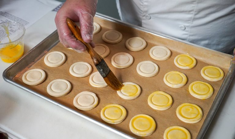 A baker wearing plastic gloves uses a pastry brush to apply a golden egg wash onto rows of small, circular, unbaked pastries arranged on a parchment-lined baking sheet.