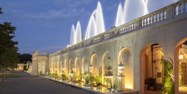 Long diagonal shot of stone facade containing multiple arches, decorated with aquatic plants and lights, fronted by a long narrow pool, topped by a stone railing and soaring fountains against a sky at dusk.