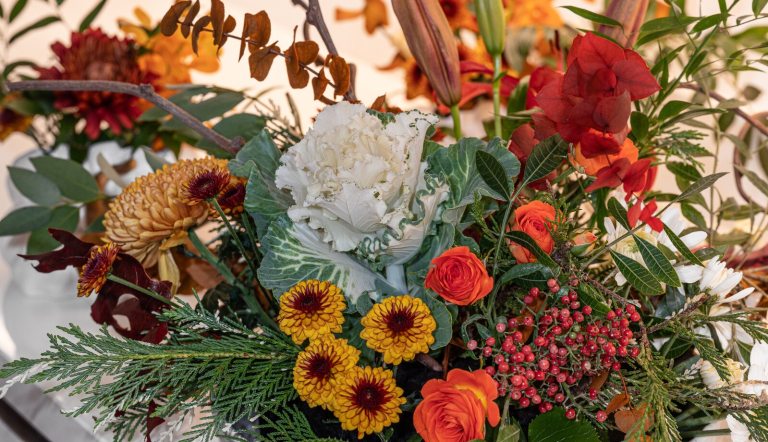A close-up of a decorative floral arrangement featuring a central white ornamental cabbage, surrounded by warm-toned orange and yellow chrysanthemums, red berries, and autumn-colored leaves.