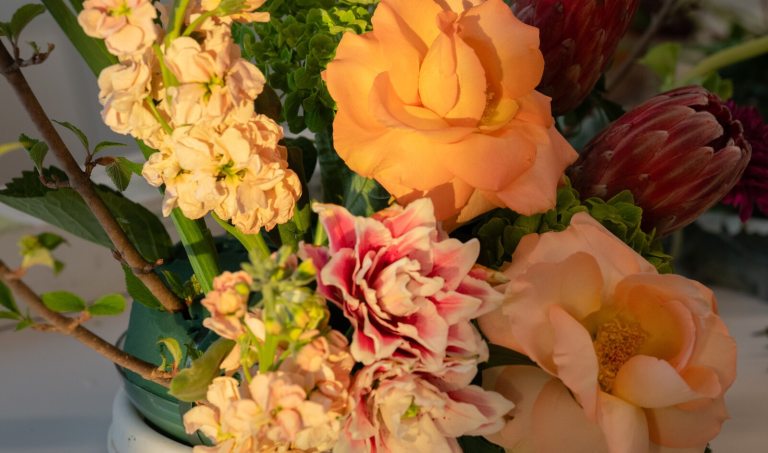 A detailed floral centerpiece showcasing peach-colored roses, pale pink stock, and deep red protea blooms in a green floral foam base.