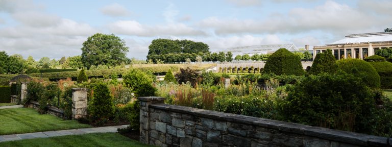 Long view of stone wall bordering an outdoor garden, with conservatory in the background.