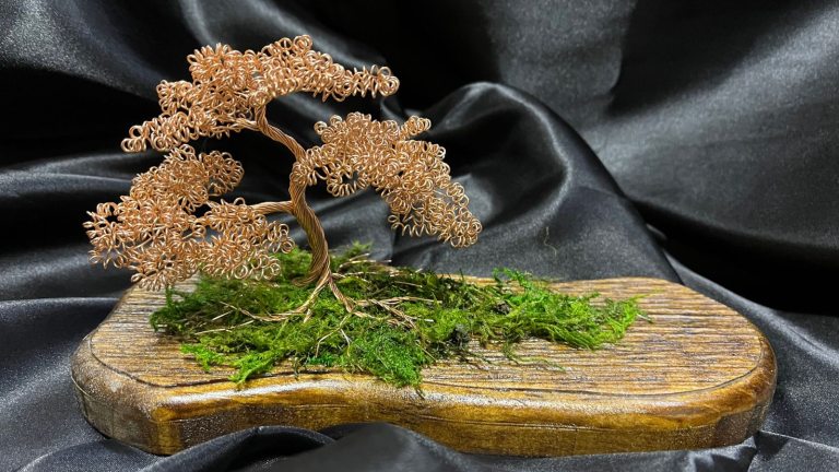 A wire bonsai sculpture on a wooden board sitting against a black fabric backdrop.