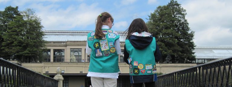 Two scouts with badges on their uniform vests face a large conservatory, with their backs to the camera.