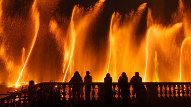 Guests in black silhouette stand at a rail watching orange fountains rising like flames.