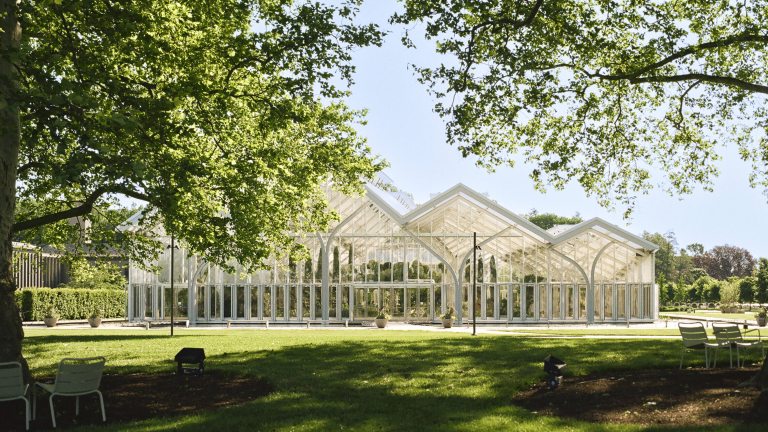 Large trees shade a green lawn in front of a large glass house with peaked arches.