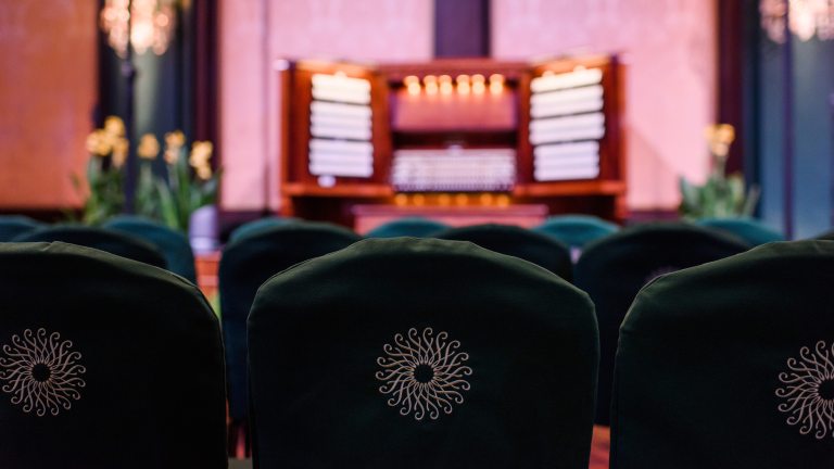 Images of empty green chairs with the Longwood logo on back, awaiting a performance, with a blurred image of an organ console in the background.