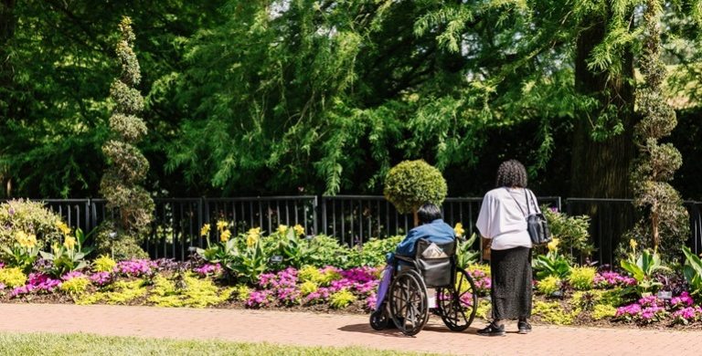 Two people, one standing, and one in a wheelchair, admiring colorful garden beds. 