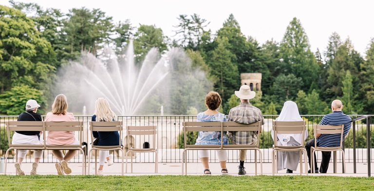 People seated outdoors in tan chairs, watching fountains in front of them. 