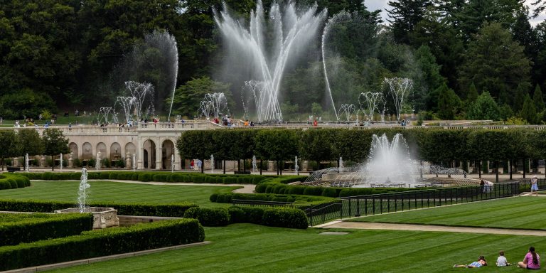 Guests enjoy the dance of fountains in various shapes and sizes in a deep green garden with a stone facade.